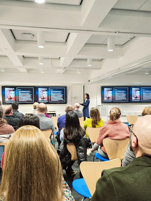 A view the back of a room, where Dr Ciorsdaidh Watts is presenting on AI ethics. She's standing beside two projector screens and gesturing towards them. 