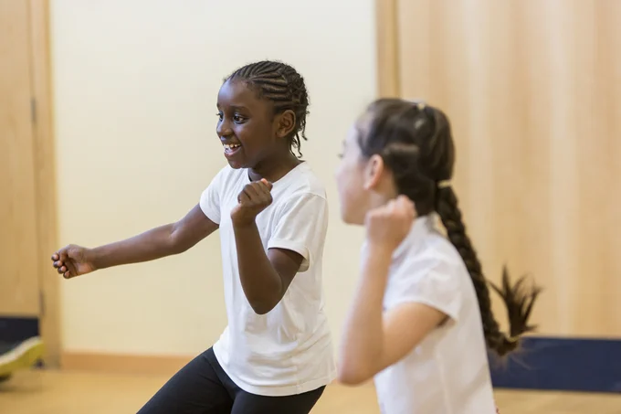 Two smiling children are engaged in a lively dance. The child on the left is wearing a white t-shirt and black pants, expressing happiness with raised arms. The child on the right, also in a white shirt, has their hair in braids and mirrors the joyful movement. The background features a light wooden wall, enhancing the energetic atmosphere of the scene.