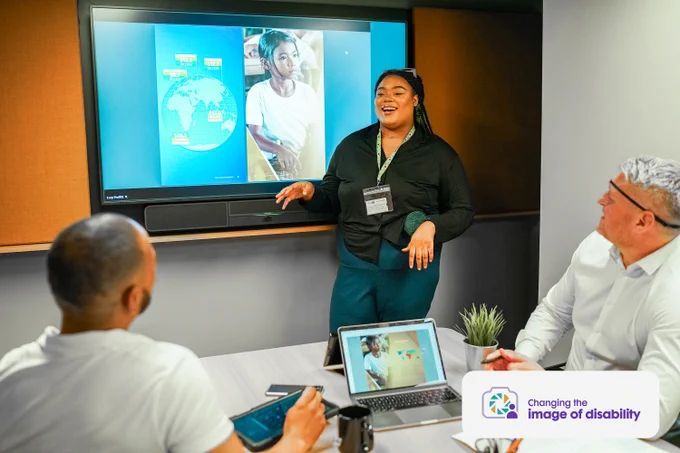 Professional wearing a sunflower lanyard gives a presentation to 2 colleagues in a meeting room. The person on the far right is wearing an eye-patch. The Business Disability Forum 'Changing the image of disability' logo is in the lower right corner of the image.