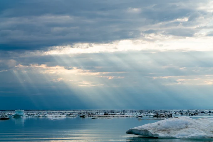 Rays from the sun poke down through the thick clouds hovering above the ocean below. Large pieces of sea ice float in the calm sea waters. 
