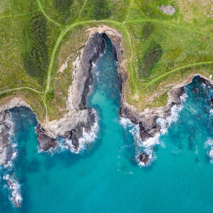 Aerial view the Cornish coastline, it shows vibrant turquoise waters with rocky cliffs and green grassy terrain.