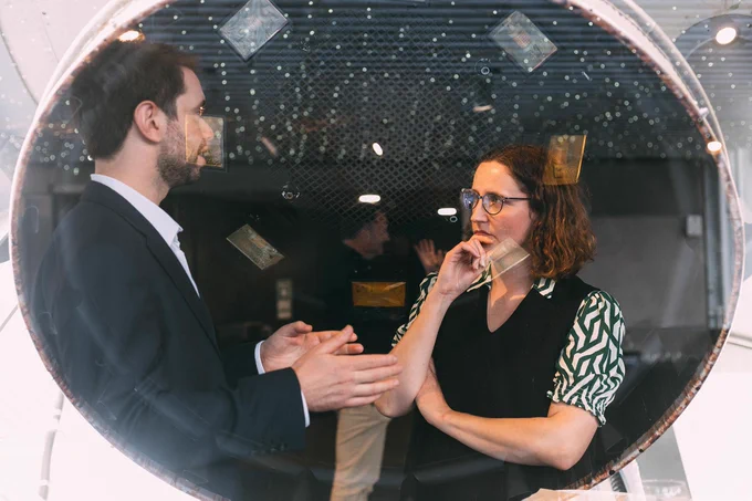 Two individuals are engaged in a conversation at our Leicester Innovate event last year. There are circular windows, they are wearing professional attire. One is in a black suit and the other in a black and green patterned shirt. Small notes are visible on the window.