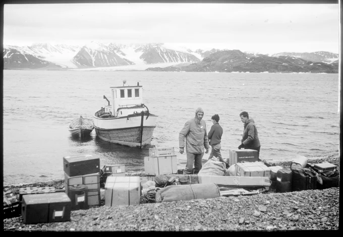 A group of 3 men unloading equipment in boxes and bags from a small boat on to a stony shore. The mountain range in the background is covered in snow.