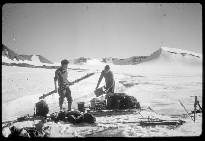 Two men tending to sleds with equipment in a snowy landscape.
