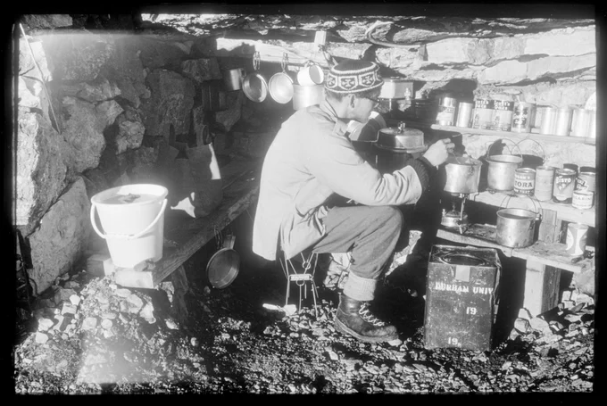 A man well wrapped against the cold tending a pan in a makeshift kitchen with shelves made from planks of wood.