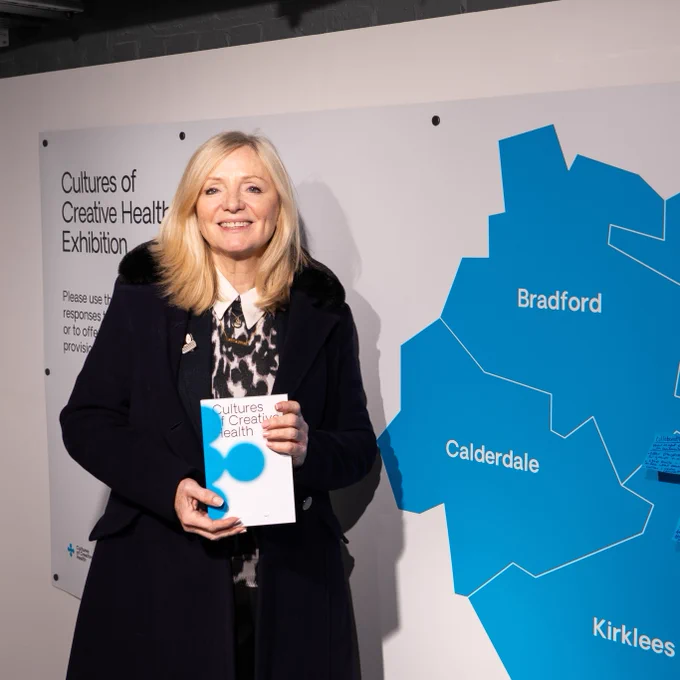 Mayor of West Yorkshire, Tracy Brabin, holding a booklet that says "Cultures of Creative Health" on the front. She is stood in front of a large blue map of West Yorkshire.