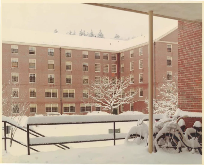 Vintage photo of a snow-covered campus, featuring a large brick building with multiple windows, a tree with snow-laden branches, and snow-covered bikes in the foreground.