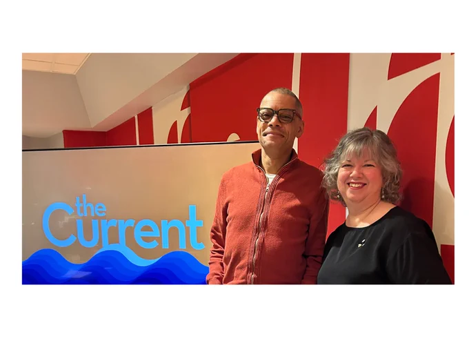 A man and a woman are smiling indoors. To the left is a monitor that displays "the Current" in blue text above a blue stylized wave.