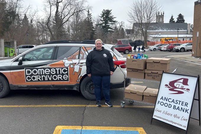 Photo of a person from Domestic Carnivore standing with a cart of food donations and the Ancaster Food Bank sandwich board sign.