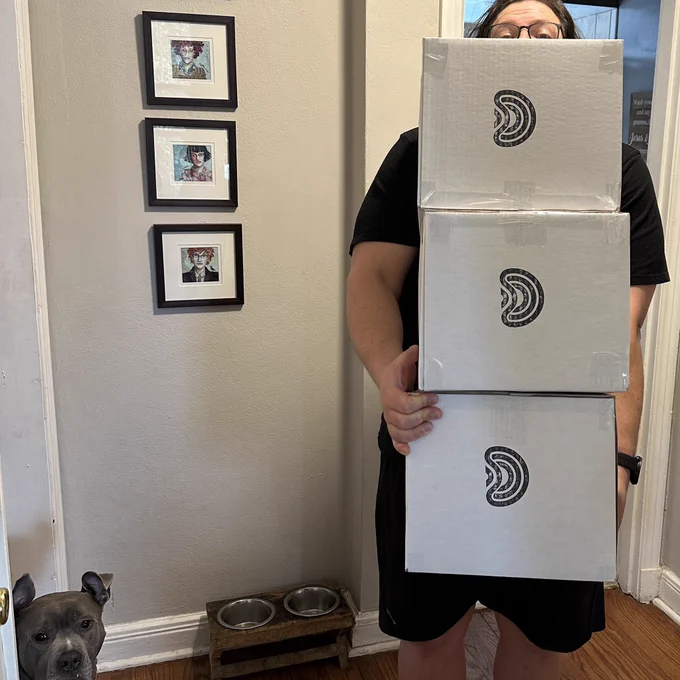Waldo holding three boxes of FDMS8 Print-it-Yourself kits, his dog Estella is peeking around the corner, and Waldo is peeking over the boxes