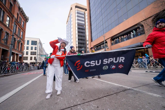 C S U’s Wiz Kid smiles and holds a banner in one hand while gesturing the C S U RAM horns with the other hand while walking in the National Western Stock Show Kick-Off Parade. The banner reads “C S U Spur.”