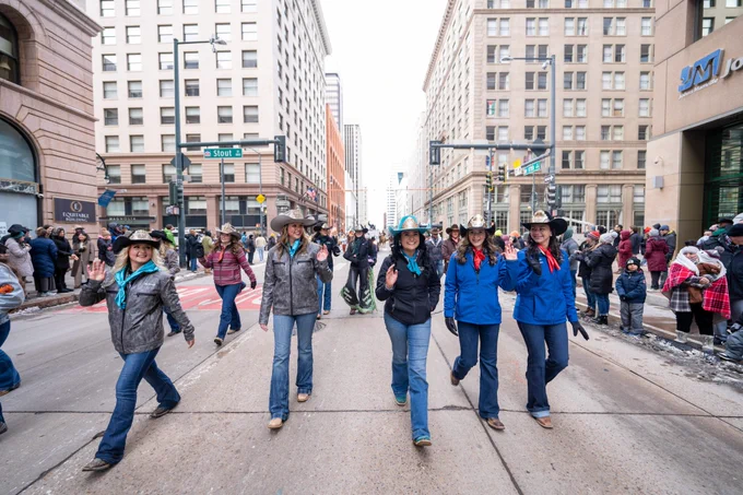 A group of people adorned in boots, cowboy hats, and bandanas smile while waving to the crowd at the National Western Stock Show Kick-Off Parade.