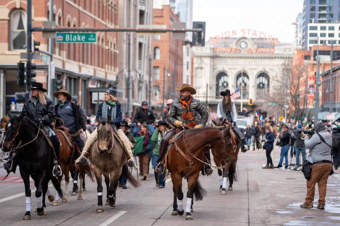 A group of people riding horseback at the National Western Stock Show Kick-Off Parade strut down 17th Street in downtown Denver, while Union Station stands tall in the background on a cold winter day. 