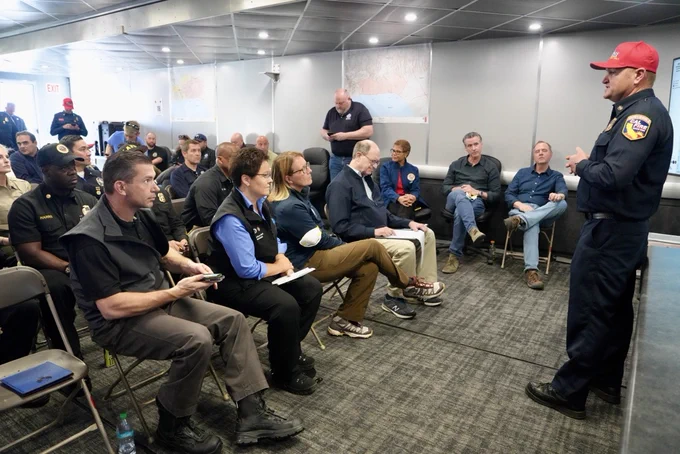 A group of people sit and listen to someone standing at the front of a room speaking to them.