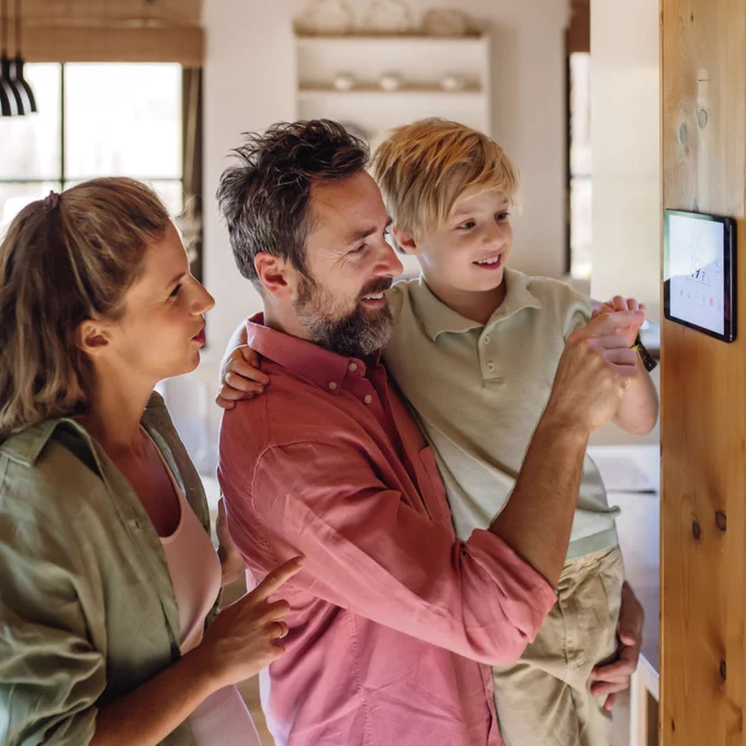 Dad holds his son up to change the settings on a smart thermostat on the wall. Mom smiles standing behind them.