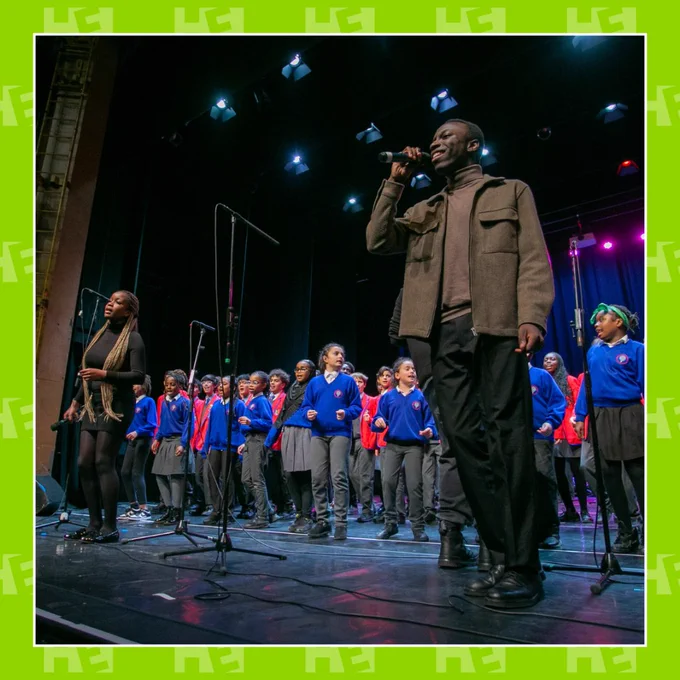 A group of young people and two adults are standing on stage performing their rehearsed songs. this image is in a green Hackney Empire border. 