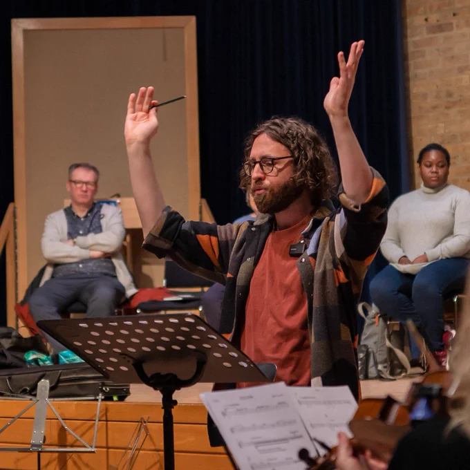 Photo of composer Gavin Higgins conducting at the MidWinter Composers course. His arms are raised above his head and two of the composers are seated on a stage behind him.