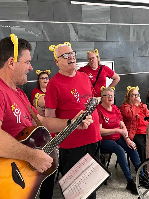 A choir in red t-shirts are singing, with some people standing and some people sitting. One person plays a guitar and has a music stand in front of them with sheet music displayed. 