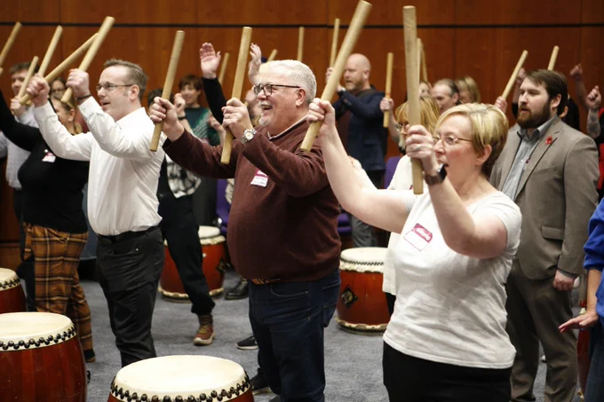 Rows of delegates at the Gathering take part in a Taiko drumming workshop. Everyone has their hands raised high holding drum sticks, with a drum sat in front of them while they stand. 