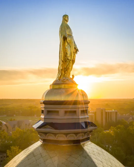 Close-up of a golden statue of the Virgin Mary on top of the Golden Dome at the University of Notre Dame, illuminated by a golden sunrise and campus in the back.