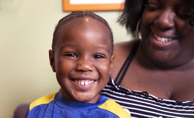 A smiling child wearing a blue and yellow shirt, accompanied by a smiling adult in a striped top, in a warmly lit room.