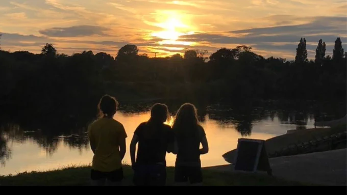 Three young people by a river, watching a sunset with trees and clouds in the background.