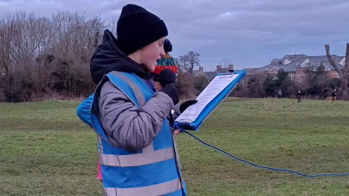 Young person in a hi-vis vest and winter clothing holding a microphone and reading from a clipboard in a park setting.