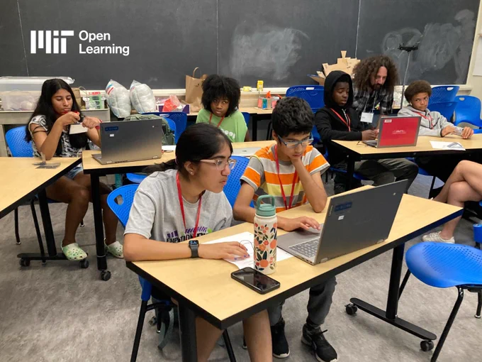 High school students engaged in learning activities at a classroom. They are seated at tables with laptops and other educational materials around them. Logo for MIT Open Learning is in the corner.
