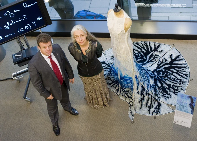 Helen Storey stands beside a man next to a mannequin showcasing an artistic white and blue gown with intricate designs, displayed over a circular carpet that complements the dress's patterns. A large screen with handwritten text and an information board are visible in the modern interior setting.