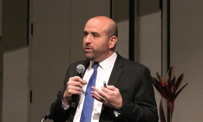 Rick: a white man of middle years in a black suit with white shirt and blue tie, speaking into a microphone and holding a usb drive