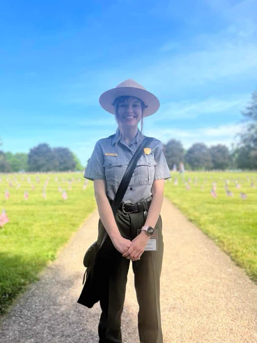 A NPS ranger stands on path with green grass and flags in the background. Blue sky. 