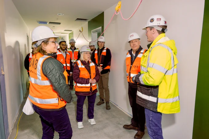 Senator Murray is standing, wearing an orange safety vest and white hard helmet, with her hands folded together. She is standing with at least six other people pictured who are all looking at a man in a bright yellow safety jacket who is speaking. They are in a white and green room with a pink wire above them that leads across the ceiling.