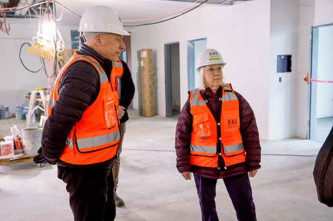 Senator Murray is standing in a jacket, orange safety vest, and white hard helmet, looking off to the right of the camera. To the left of the Senator, a man wearing the same thing is standing with his hands behind his back. They are in a white room with concrete floors and behind them there is a large light with wires coming out of the ceiling and construction equipment including paint and caution tape.