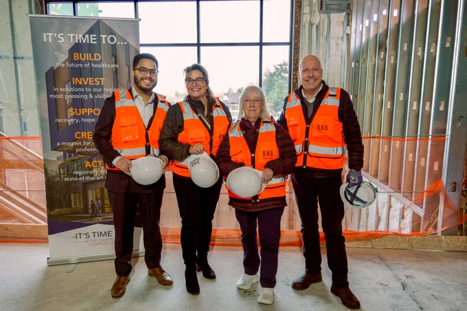 Senator Murray is standing with three other people, all of which are wearing orange safety vests, carrying hard helmets, and smiling at the camera. They are standing in a large building that is still under construction. Behind them is an orange safety net and a sign that shows the what the building will ultimately look like once construction is completed.