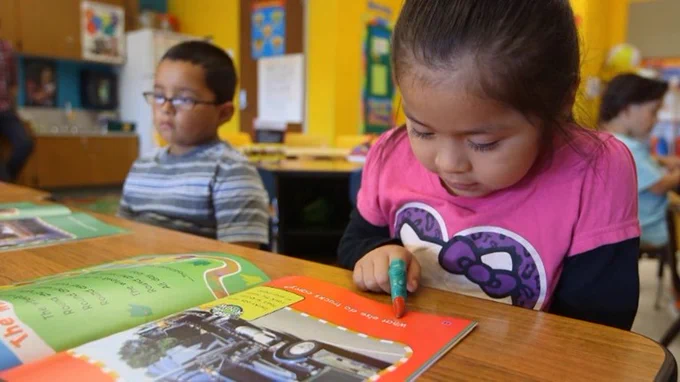 wo young students sit at a desk in a classroom working on their reading skills.