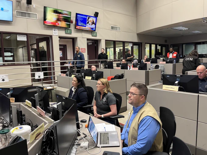 Inside a busy emergency operations center, multiple professionals are seated at desks equipped with computers and monitors, intently managing a situation. There are digital screens displaying news and updates in the background.