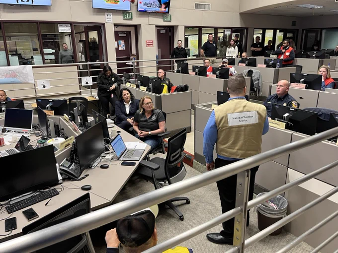 Emergency operations center with multiple desks and computer stations. Individuals are seated and engaged in various tasks, some interacting with one another, and a person wearing a 'National Weather Service' vest stands addressing the group. Monitors display information around the room.