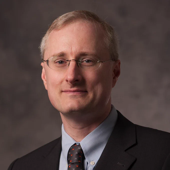 Portrait of Juergen Hahn wearing glasses, a dark blazer, and a tie with a subtle pattern, set against a textured gray background.
