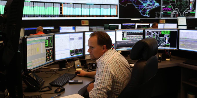 A person working at a desk in a control room with multiple computer monitors displaying data and graphs.