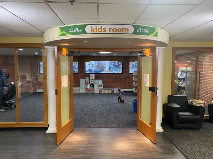 The Kids Room in the Lobby. Doors stand open with a sign that reads “kids room (it’s just temporary!)" Some kids books, toys and seating are available. A toddler toddles by.