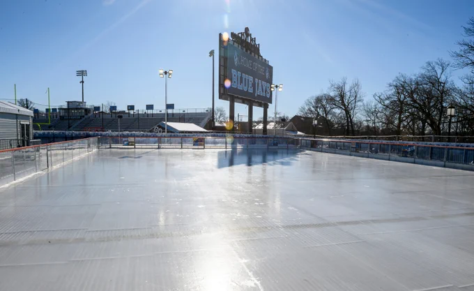 A sunlit outdoor ice skating rink with a large scoreboard in the background.