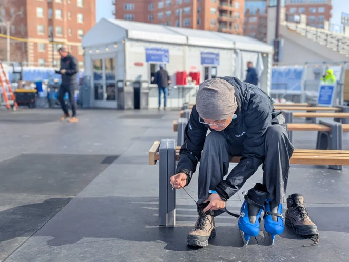 A person dressed in winter sportswear is adjusting the strap on their shoe and at an outdoor ice skating rink.