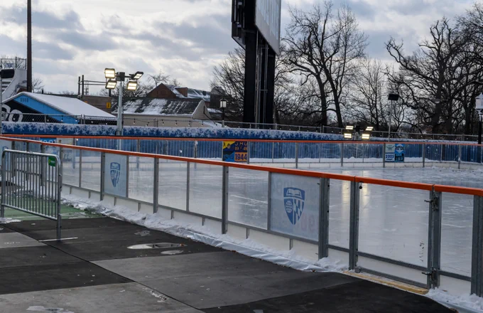 Outdoor ice skating rink with surrounding barriers displaying the John Hopkins University shield, set against a backdrop of leafless trees and a cloudy sky.