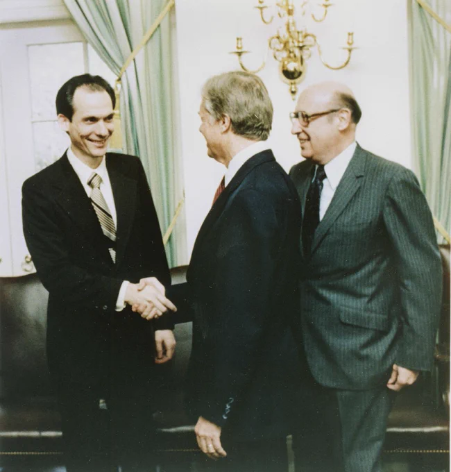 An archival photo from 1977 shows three men dressed in suits. The man at the left is Voyager project scientist Ed Stone. He is dark-haired and smiling as he shakes the hand of the man in the middle, President Jimmy Carter. Carter faces away from the camera. At right, a third man in a pinstripe suit and glasses smiles toward the men shaking hands.