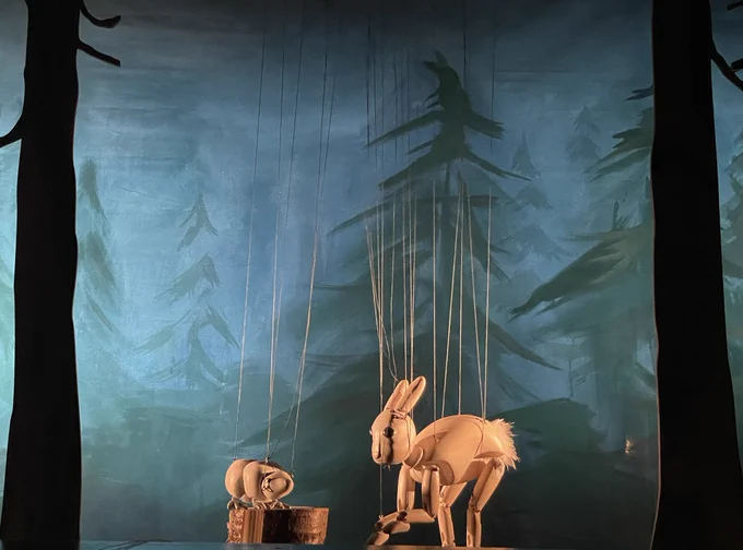 A snow hare wooden marionette is talking to a lemming wooden marionette who’s standing on a log against a painted green forest background.