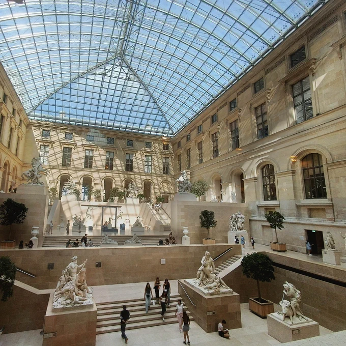 Entrada al Museo del Louvre. Techo de cristal en una gran sala con escalinatas y estatuas. Punto de vista elevado.