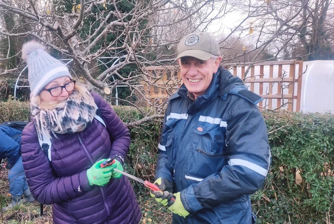 A man holding a pruning saw stands next to a smiling woman in an orchard. 