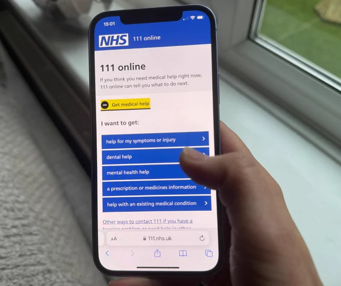A hand holding a smartphone displaying the NHS 111 online webpage with options for medical help.