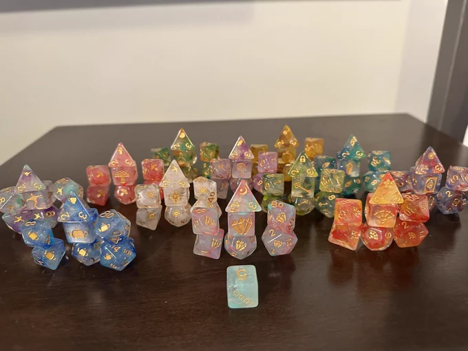 Twelve sets of colorful dice displayed on a wooden table with a d6 featuring the Critit logo in front of them