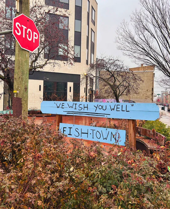 A blue sign in the Fishtown Rose Garden on a rainy day. It reads: We Wish You Well Fishtown.